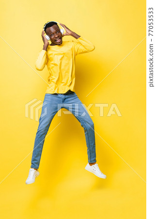 Happy young African man wearing headphones listening to music and jumping Happy young African man wearing headphones listening to music and jumping 77053533