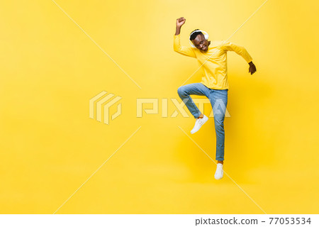 Happy energetic young African man wearing headphones listening to music and jumping with hand up Happy energetic young African man wearing headphones listening to music and jumping with hand up 77053534