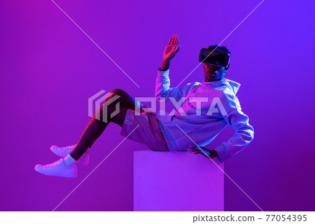 Young African man wearing VR glass headset in futuristic purple cyberpunk neon light background 77054395