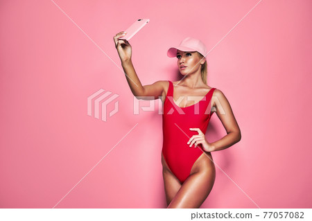 Smiling young blonde woman in red one-piece swimsuit cap doing selfie shot isolated on pink background. Smiling young blonde woman in red one-piece swimsuit cap doing selfie shot isolated on pink background. 77057082