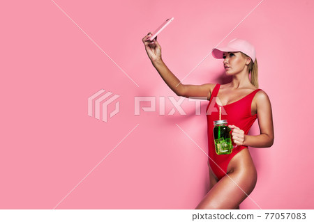 Smiling young blonde woman in red one-piece swimsuit cap doing selfie shot isolated on pink background. Smiling young blonde woman in red one-piece swimsuit cap doing selfie shot isolated on pink background. 77057083
