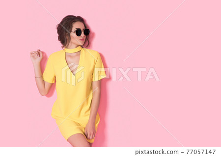 Beautiful girl in yellow dress wearing sunglasses posing, smiling on pink background in studio.  77057147