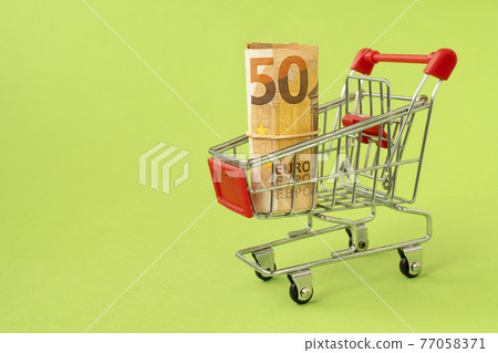 Shopping cart with roll of Euro money on green background Shopping cart with roll of Euro money on green background 77058371