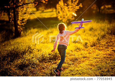 Little girl launches a toy plane into the air in the park outdoor. Child launches a toy plane. Beautiful little girl stands on the grass and launches a pink toy plane. 77058945
