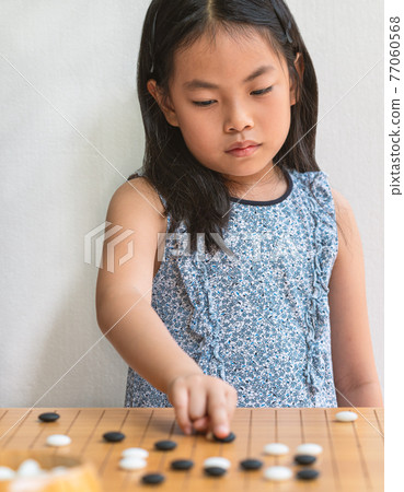 Portrait Asian child girl plays Go, the Japanese board game or Chinese board game, focus on the board game, white wall, vertical image. 77060568