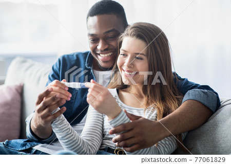 Happy young interracial couple holding pregnancy test, looking at positive result Happy young interracial couple holding pregnancy test, looking at positive result 77061829