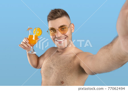Smiling millennial guy in sunglasses holding cool orange cocktail and taking selfie on blue studio background Smiling millennial guy in sunglasses holding cool orange cocktail and taking selfie on blue studio background 77062494