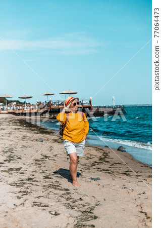 Adult blond woman with straw summer hat and yellow cotton shirt walks along the beach on a sunny day. Vertical photo orientation Adult blond woman with straw summer hat and yellow cotton shirt walks along the beach on a sunny day. Vertical photo orientation 77064473