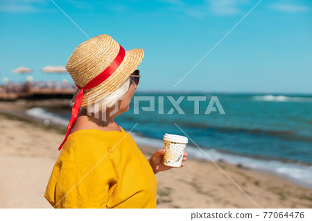 Blonde woman holding a paper cup with coffee to go while standing by the sea. Woman in a straw hat walks and enjoys a beautiful view 77064476