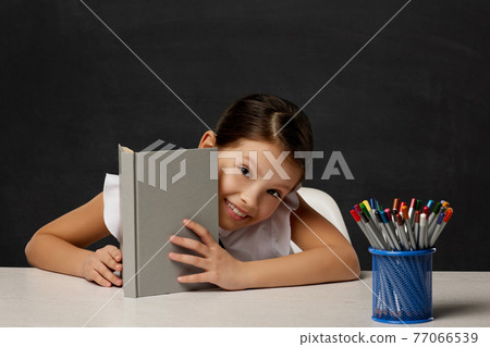 little schoolgirl looks out from behind the book little schoolgirl looks out from behind the book 77066539