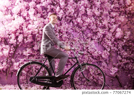 Side view of smiling bridegroom riding bicycle against wall covered with pink flowers 77067274