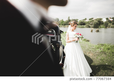 Beautiful newlywed couple posing near retro black car Beautiful newlywed couple posing near retro black car 77067384