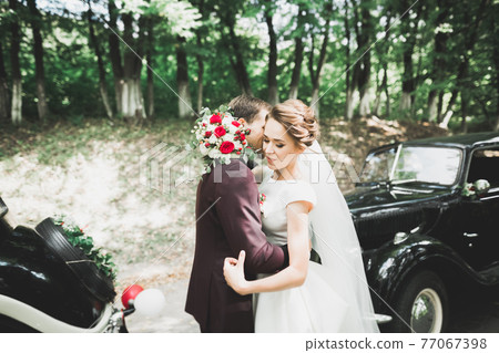 Beautiful newlywed couple posing near retro black car Beautiful newlywed couple posing near retro black car 77067398