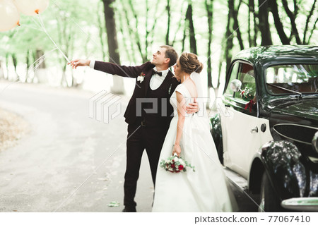 Beautiful newlywed couple posing near retro black car Beautiful newlywed couple posing near retro black car 77067410