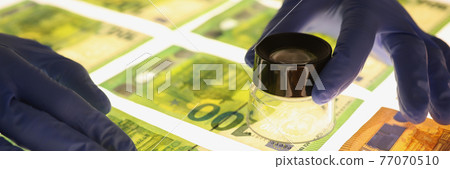 Hands in rubber gloves holding magnifying glass over banknotes closeup 77070510