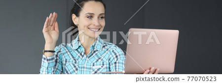 Young woman holding laptop and waving hand Young woman holding laptop and waving hand 77070523
