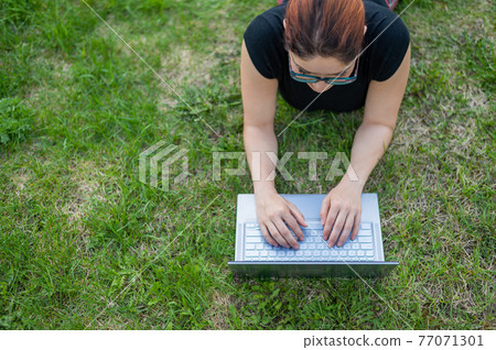 Mobile office. Top view of a freelancer woman typing a laptop while lying on the lawn. Work outdoors. Girl studying on a wireless computer on the grass in the park. Mobile office. Top view of a freelancer woman typing a laptop while lying on the lawn. Work outdoors. Girl studying on a wireless computer on the grass in the park. 77071301