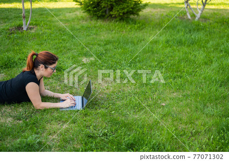 Red-haired woman on the lawn in the park and types on the laptop Red-haired woman on the lawn in the park and types on the laptop 77071302