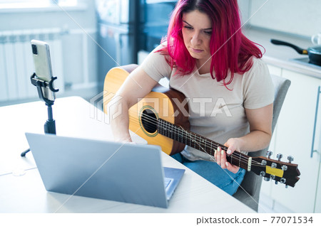 A woman sits in the kitchen during a remote acoustic guitar lesson. A girl learns to play the guitar and watches educational videos on a laptop A woman sits in the kitchen during a remote acoustic guitar lesson. A girl learns to play the guitar and watches educational videos on a laptop 77071534