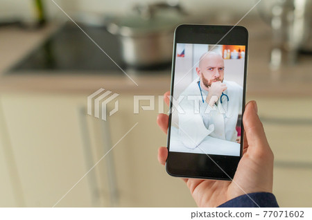 A serious bearded doctor communicates with a patient via a video call on a smartphone. Online medicine concept. Woman in the kitchen takes therapist remote consultation on mobile. A serious bearded doctor communicates with a patient via a video call on a smartphone. Online medicine concept. Woman in the kitchen takes therapist remote consultation on mobile. 77071602
