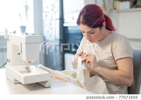 Caucasian woman sews while sitting in the kitchen. Home hobby Caucasian woman sews while sitting in the kitchen. Home hobby 77071608