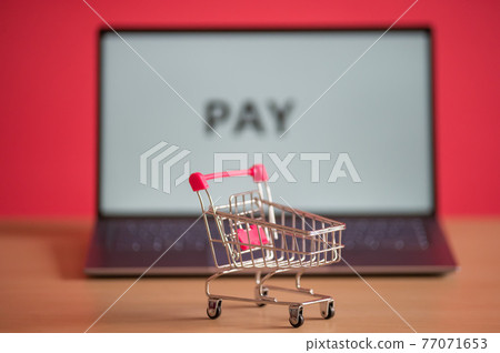 Mini shopping trolley stands in front of a laptop on a pink background. Online shopping concept. The word pay on a computer screen. Remote purchase of products and things on the Internet. Mini shopping trolley stands in front of a laptop on a pink background. Online shopping concept. The word pay on a computer screen. Remote purchase of products and things on the Internet. 77071653