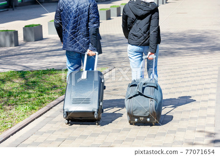 Two people, a man and a woman, walk along the cobbled sidewalk, carrying a travel bag and a suitcase on wheels behind them. 77071654