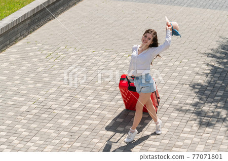 Happy caucasian young woman in hat and shorts holding a large red suitcase at an open age 77071801