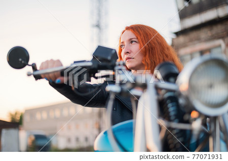 Curly red-haired woman in a black leather jacket sits on a motorcycle. Portrait of a serious girl driving a bike. 77071931