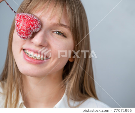 Cute woman with braces on her teeth holds a candy in the form of a heart on white background Cute woman with braces on her teeth holds a candy in the form of a heart on white background 77071996
