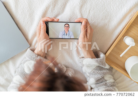 A faceless woman is at home in bed and consults with a healer using a webcam. The doctor remotely consults the patient on a video call. Medicines on a wooden tray. 77072062