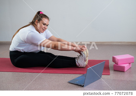 Young caucasian fat woman doing bends on a sports mat and watching a training video on a laptop. A chubby girl doing stretching remotely using video communication Young caucasian fat woman doing bends on a sports mat and watching a training video on a laptop. A chubby girl doing stretching remotely using video communication 77072078