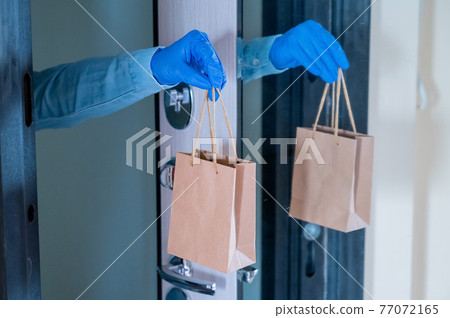 Close-up of a courier's hands in gloves with a paper bag in their hands at the front door. Quarantine delivery service. 77072165