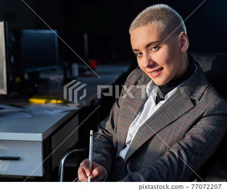Serious young woman with short hair sits in armchair in office. 77072207