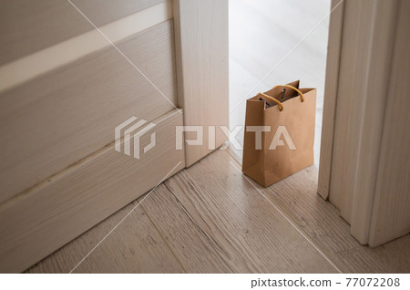 The concept of contactless delivery during the quarantine period. Kraft paper bag with online purchase at the open entrance door in case of danger of the spread of coronavirus. 77072208