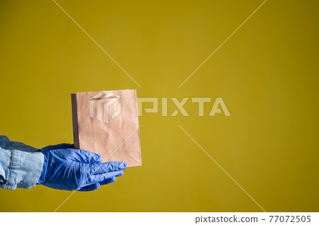 Closeup of female hands in gloves and a denim shirt. Delivery man holds an empty paper bag on a yellow background. Craft packaging for takeaway snack. Antimicrobial protection. 77072505