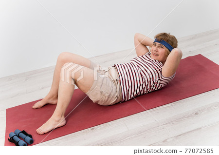 Happy old woman doing abs exercises on a sports mat. Elderly lady doing fitness. 77072585