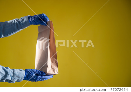 Closeup of female hands in gloves and a denim shirt. Delivery man passes an empty paper bag on a yellow background. Craft packaging for takeaway snack. Antimicrobial protection. Closeup of female hands in gloves and a denim shirt. Delivery man passes an empty paper bag on a yellow background. Craft packaging for takeaway snack. Antimicrobial protection. 77072654