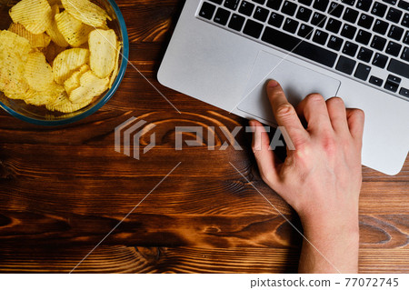 Potato chips in a glass bowl next to a laptop that controls the hand of a man. Top view. Free time. Potato chips in a glass bowl next to a laptop that controls the hand of a man. Top view. Free time. 77072745