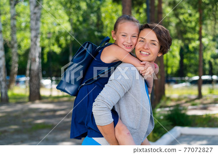 A beautiful caucasian woman stands outdoors and holds daughter on her back. A cheerful smiling girl sits on her mother back while walking in the park. Friendly loving family. 77072827