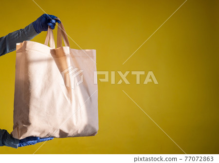 Sending eco-friendly reusable cotton bag on a yellow background. The courier holds a canvas fabric bag without plastic packaging on a yellow background. Safe delivery to the epidemic. Sending eco-friendly reusable cotton bag on a yellow background. The courier holds a canvas fabric bag without plastic packaging on a yellow background. Safe delivery to the epidemic. 77072863