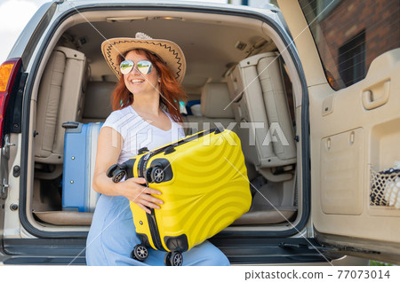 Happy red-haired woman in a cowboy hat and sunglasses going on a car trip around the country. Girl sitting in the trunk with a yellow suitcase ready for summer vacation. Independent travel. Happy red-haired woman in a cowboy hat and sunglasses going on a car trip around the country. Girl sitting in the trunk with a yellow suitcase ready for summer vacation. Independent travel. 77073014