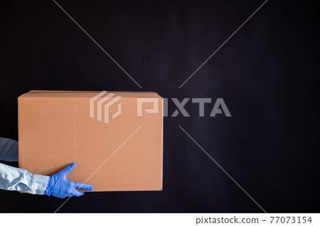 Closeup of female hands in gloves and a denim shirt. Delivery man holds a cardboard box to the customer on a black background. Antimicrobial protection in an epidemic. Closeup of female hands in gloves and a denim shirt. Delivery man holds a cardboard box to the customer on a black background. Antimicrobial protection in an epidemic. 77073154