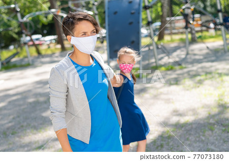 A girl in a protective mask drags her mother to the playground. Naughty moody kid wants to play. Quarantine isolation. Woman with daughter outdoors. 77073180
