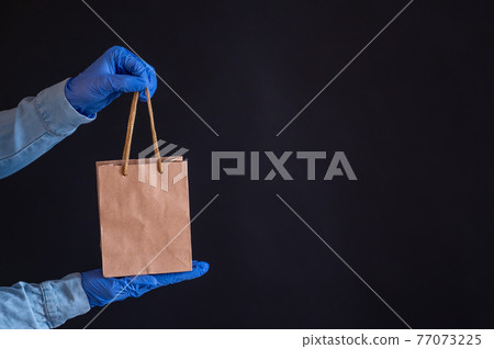 Female hands in blue gloves holds a brown paper bag with handles on a black background. Safe food delivery to your home. A courier in a denim shirt holds out a craft cardboard bag to a customer. 77073225