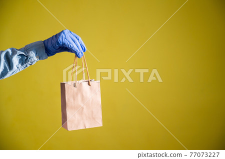 Closeup of female hands in gloves and a denim shirt. A delivery man is holding a small paper bag by the pens on a yellow background. Craft packaging for takeaway snack. Antimicrobial protection. Closeup of female hands in gloves and a denim shirt. A delivery man is holding a small paper bag by the pens on a yellow background. Craft packaging for takeaway snack. Antimicrobial protection. 77073227