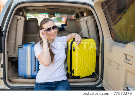 European woman in sunglasses packs her bags for a car trip. Beautiful happy girl sitting in the trunk of a car and ready for travel. Yellow suitcase with things for summer vacation. 77073279