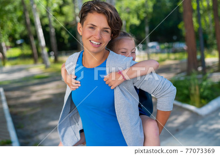 Beautiful caucasian woman holds her daughter schoolgirl on her back outdoors. A girl with a backpack lovingly hugs her mother from behind. Beautiful caucasian woman holds her daughter schoolgirl on her back outdoors. A girl with a backpack lovingly hugs her mother from behind. 77073369