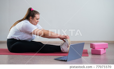 Young caucasian fat woman doing bends on a sports mat and watching a training video on a laptop. A chubby girl doing stretching remotely using video communication Young caucasian fat woman doing bends on a sports mat and watching a training video on a laptop. A chubby girl doing stretching remotely using video communication 77073370