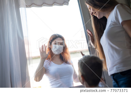 A masked woman is standing outside by a window plane, waving to her sister and nephew who are staying at home. Social distance among families in quarantine. Coronavirus. The girl at the window pane. 77073371